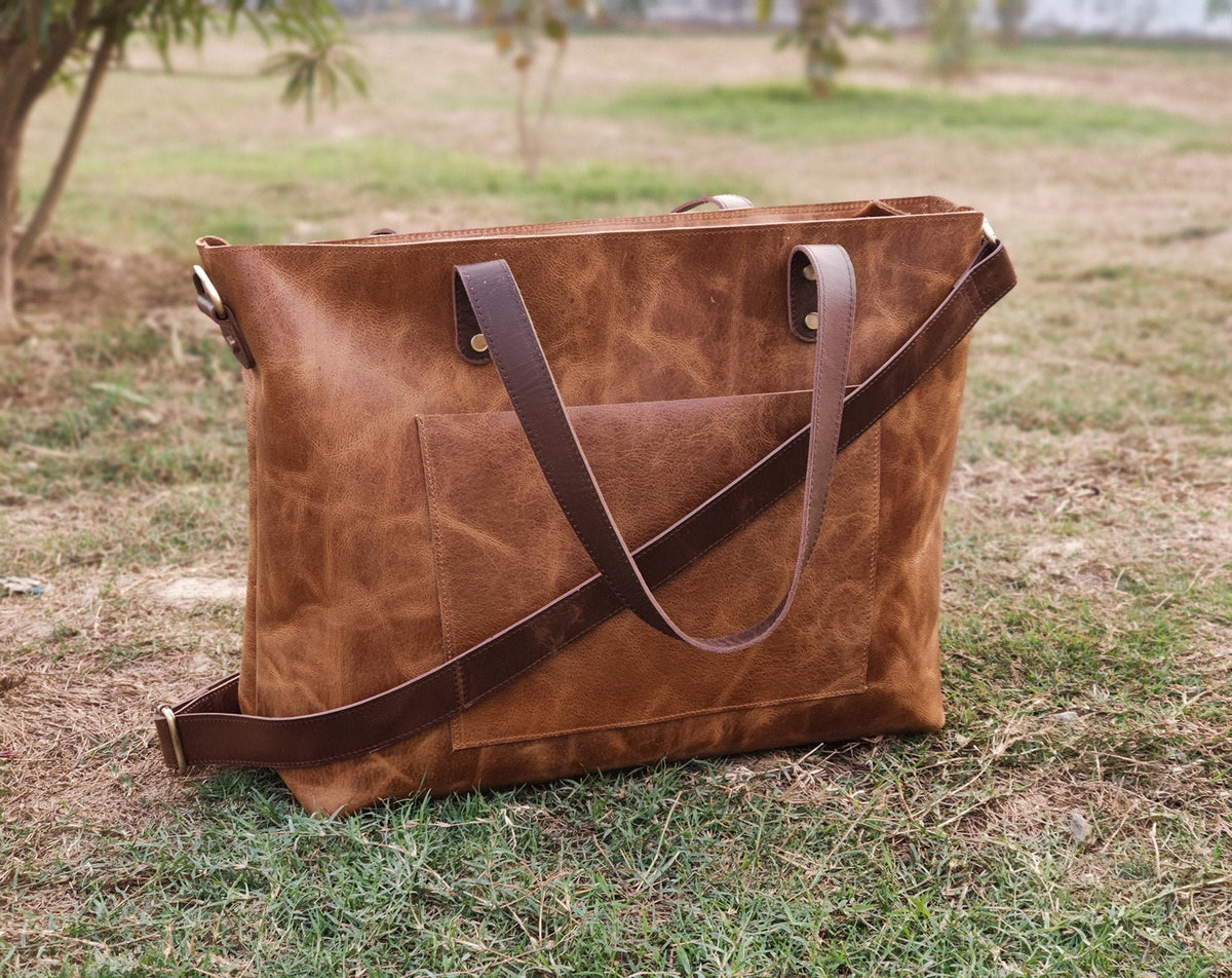 May include: A brown leather tote bag with a front pocket and a long, adjustable shoulder strap. The bag has two short handles and is sitting on grass. The leather has a distressed look.