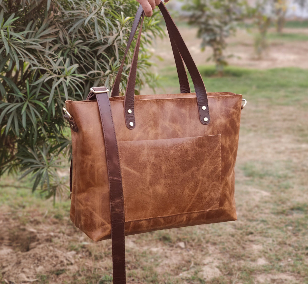 May include: A brown leather tote bag with a large pocket on the front. The bag has a long strap that can be worn over the shoulder.
