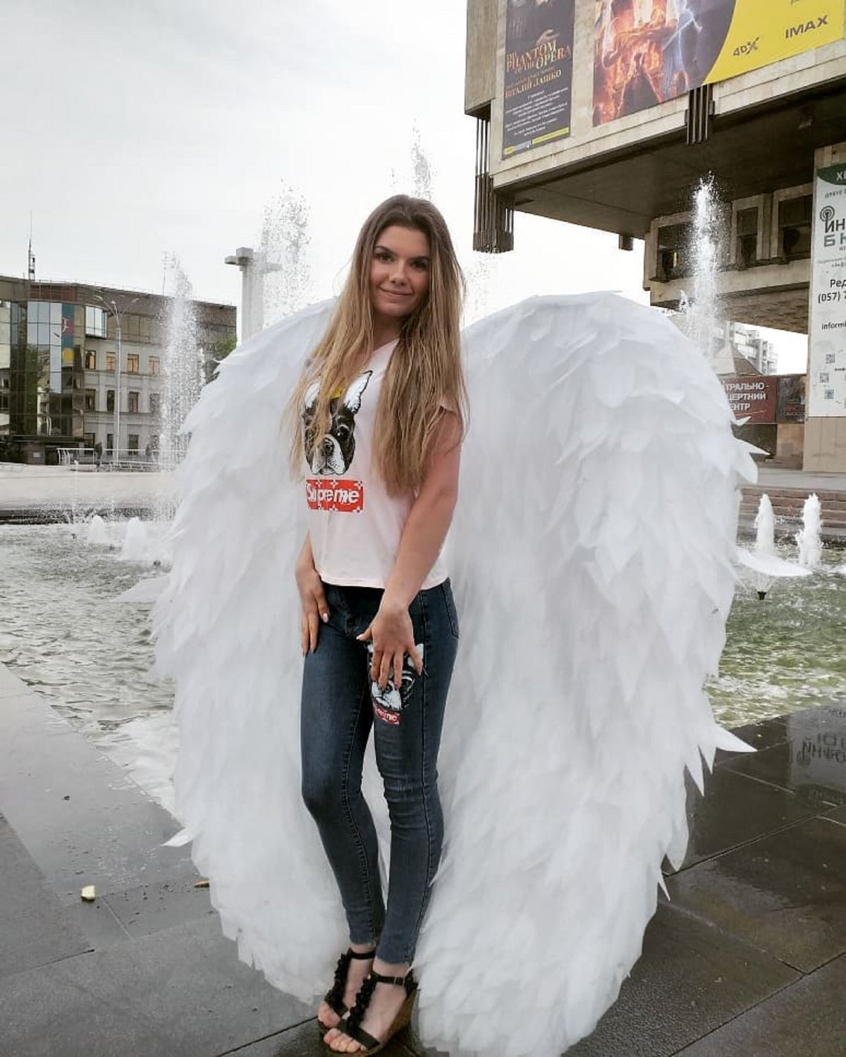 May include: A woman wearing a white t-shirt with a black and white dog graphic and the word "Supreme" printed in red. She is wearing blue jeans and white angel wings. The woman is standing in front of a fountain.