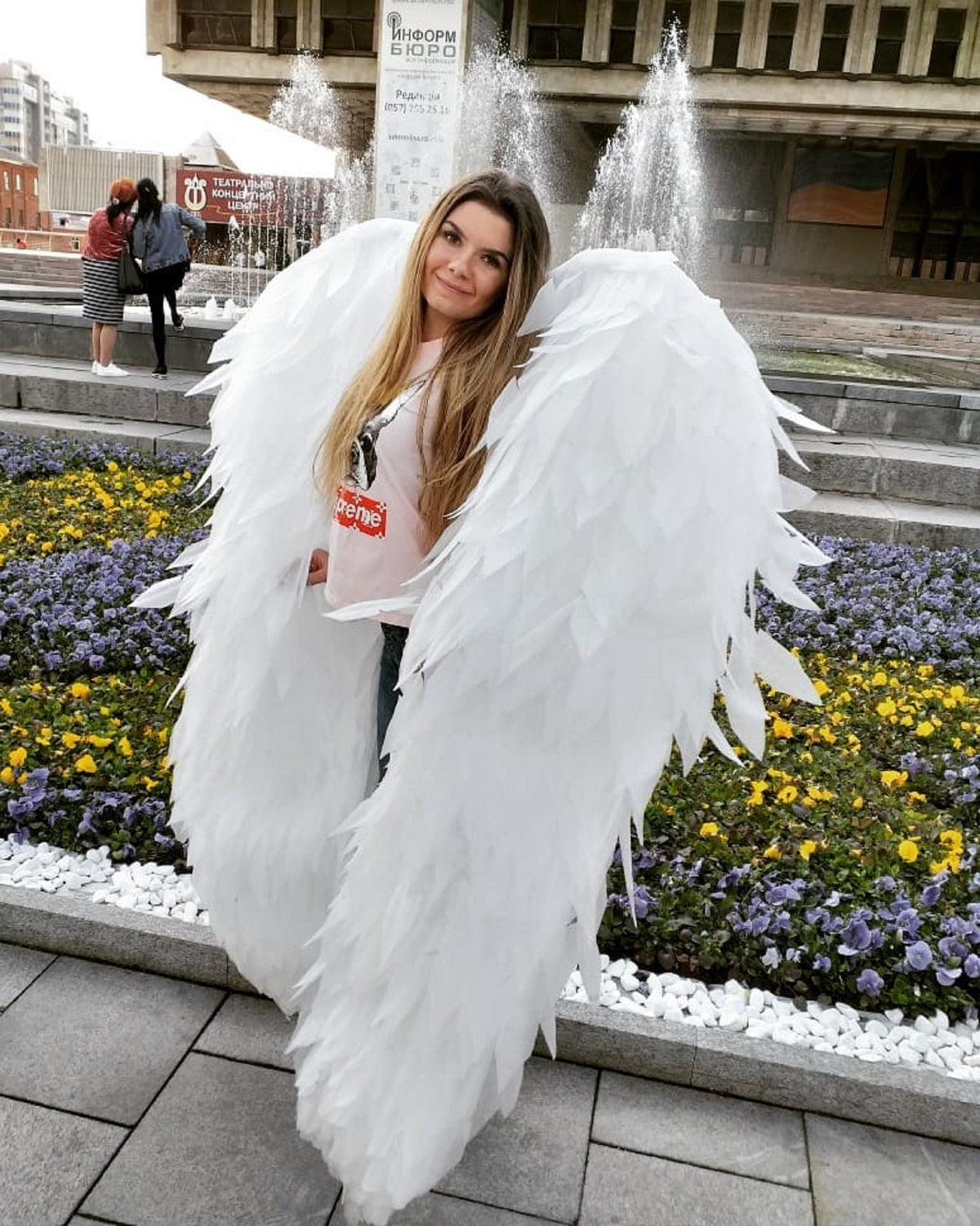 May include: A woman wearing a white t-shirt with the word "supreme" printed on it, is standing in front of a fountain with large white angel wings. The wings are made of feathers and are very detailed.