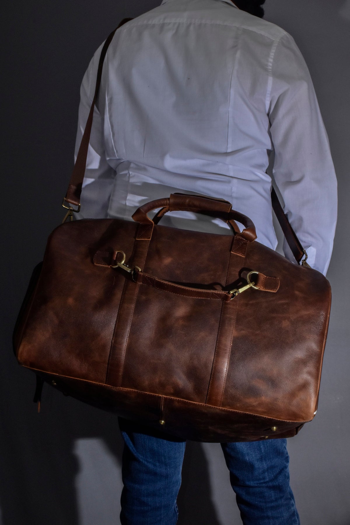 May include: A brown leather duffel bag with brass hardware and a long strap. The bag is being carried by a person wearing a white shirt and blue jeans.