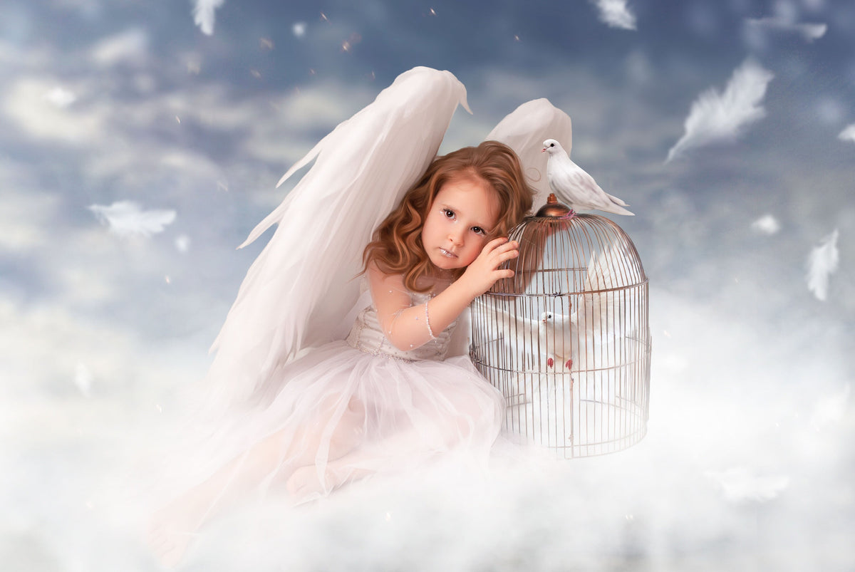 May include: A young girl dressed as an angel, wearing a white dress and large feathered wings, sits among clouds. A birdcage with a white dove inside is next to her. Another dove perches on top of the cage. The background is a sky with clouds.