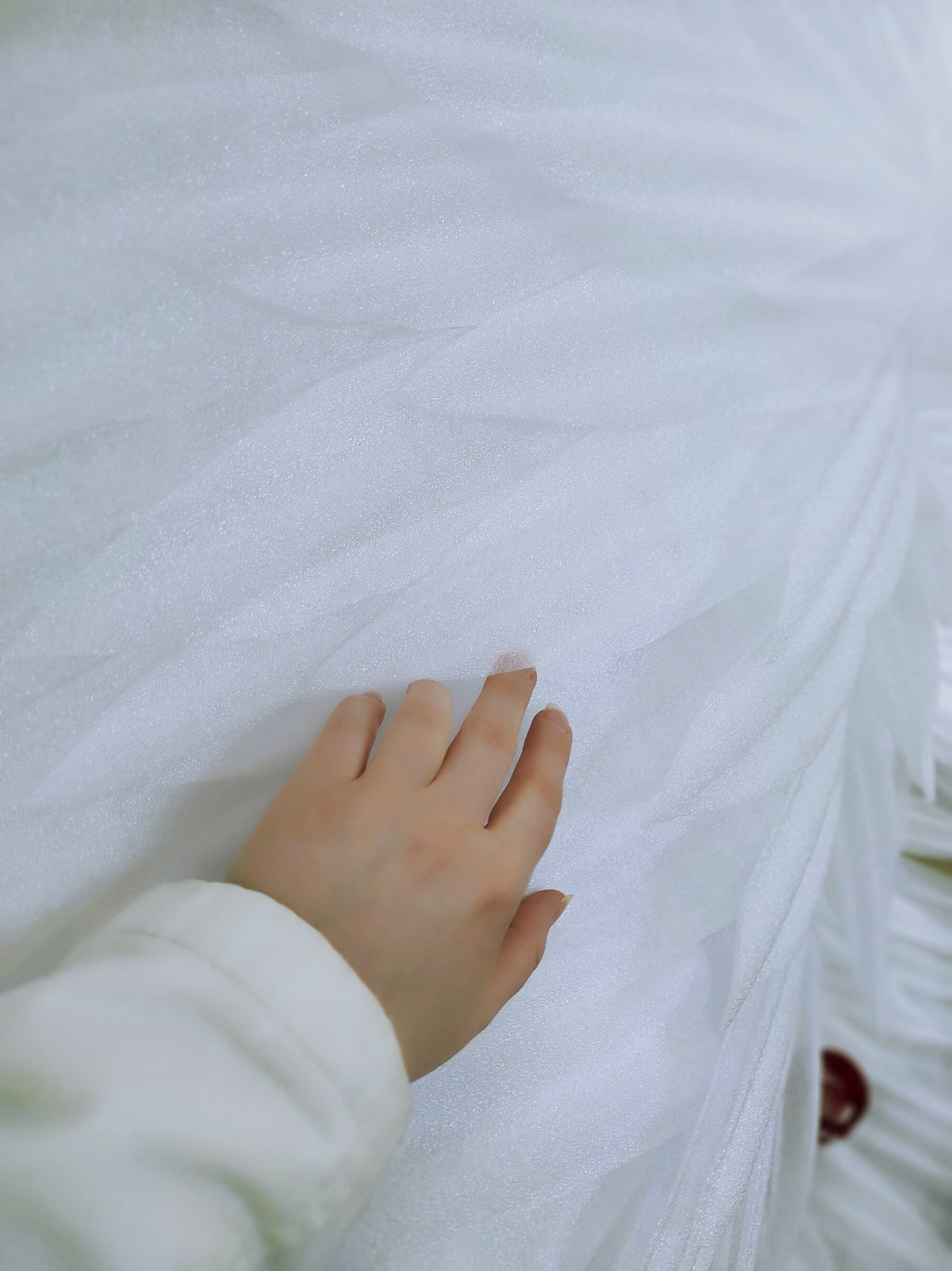 May include: Close-up of a hand touching a white, textured fabric. The fabric appears to be layered and has a soft, slightly reflective surface. The sleeve of a white garment is visible.