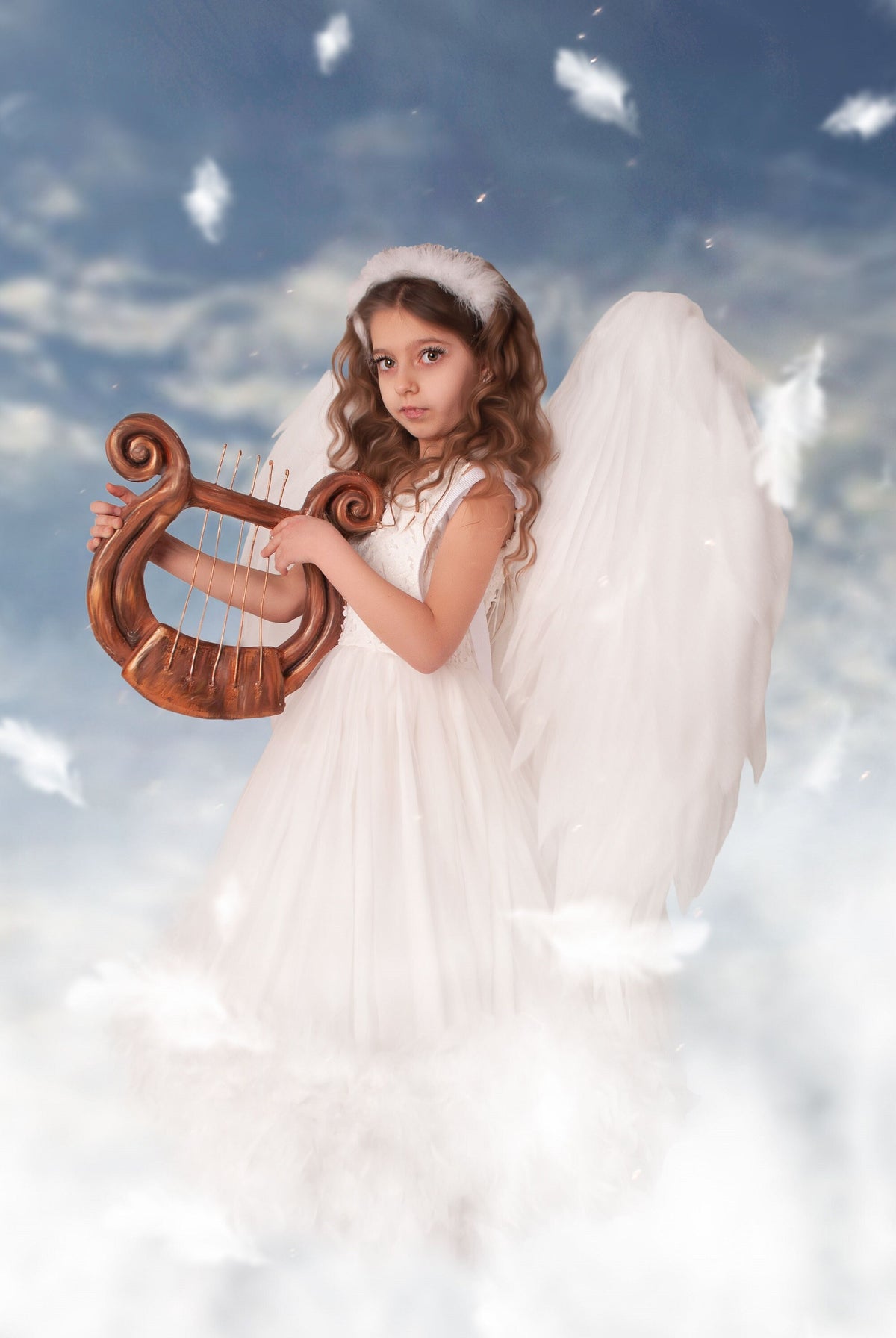 May include: A young girl dressed as an angel with white wings and a white dress plays a wooden lyre. She is standing in a cloud-like setting with white feathers floating around her.