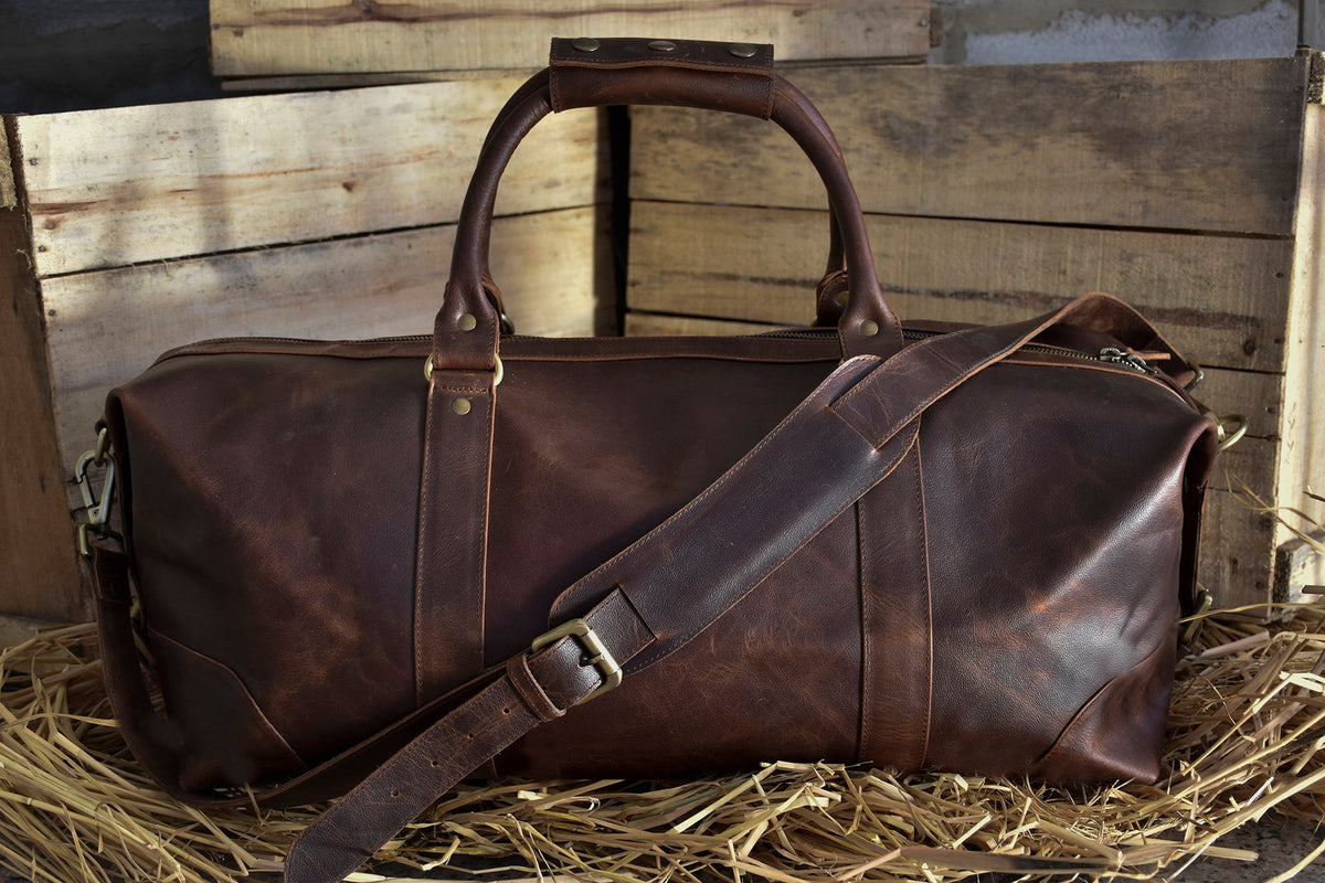 May include: A brown leather duffel bag with a long shoulder strap and two top handles. The bag has a zipper closure and brass hardware.