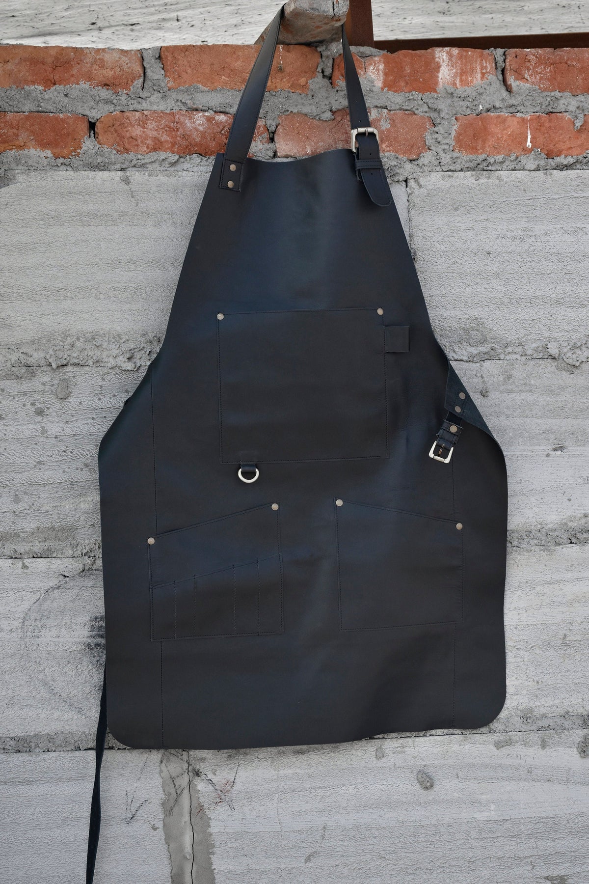 May include: A black leather apron with multiple pockets and a metal ring. The apron has adjustable straps and a buckle.
