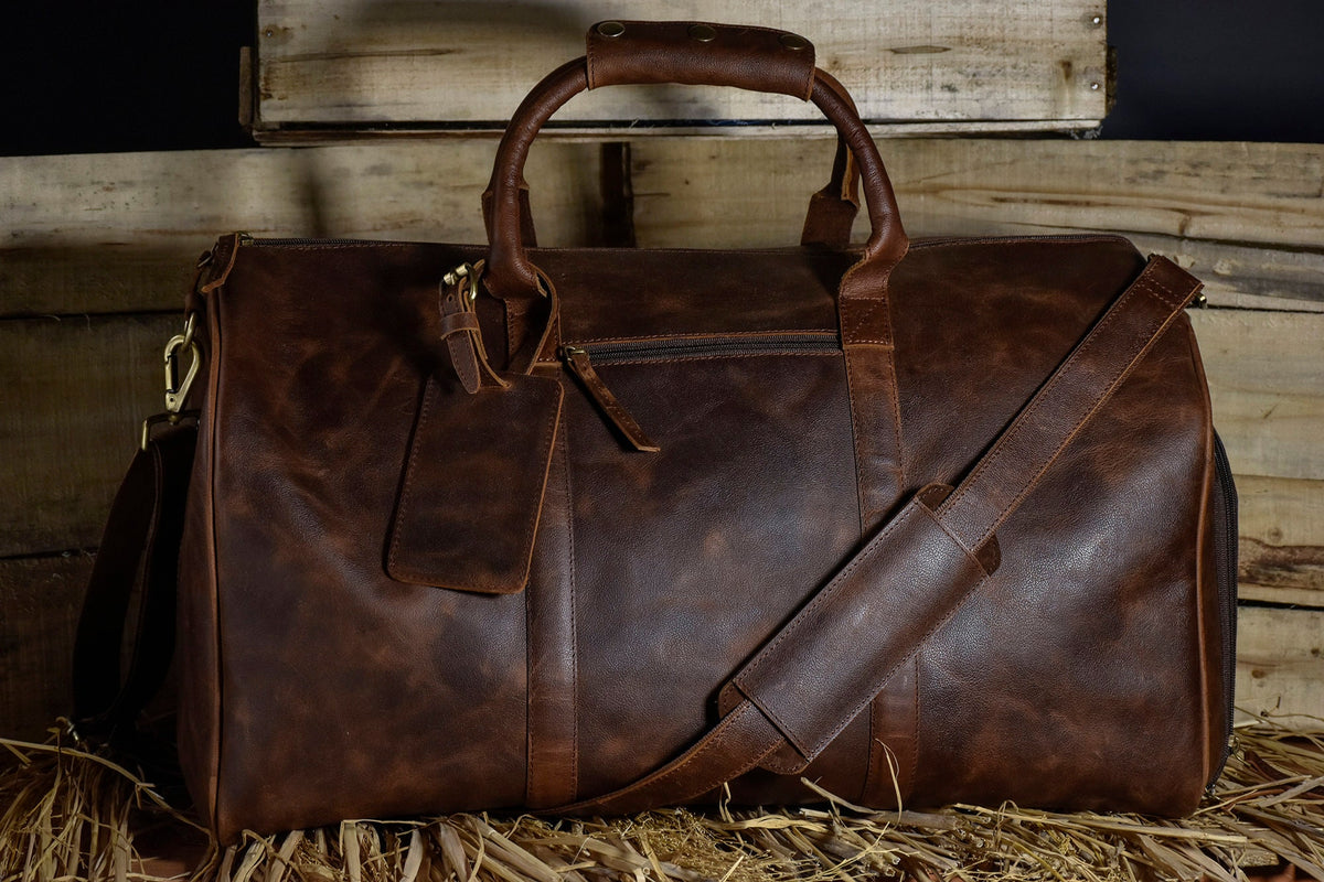 May include: A brown leather duffel bag with a top handle, shoulder strap, and a luggage tag. The bag has a zipper closure and gold-colored hardware. The leather has a distressed look.