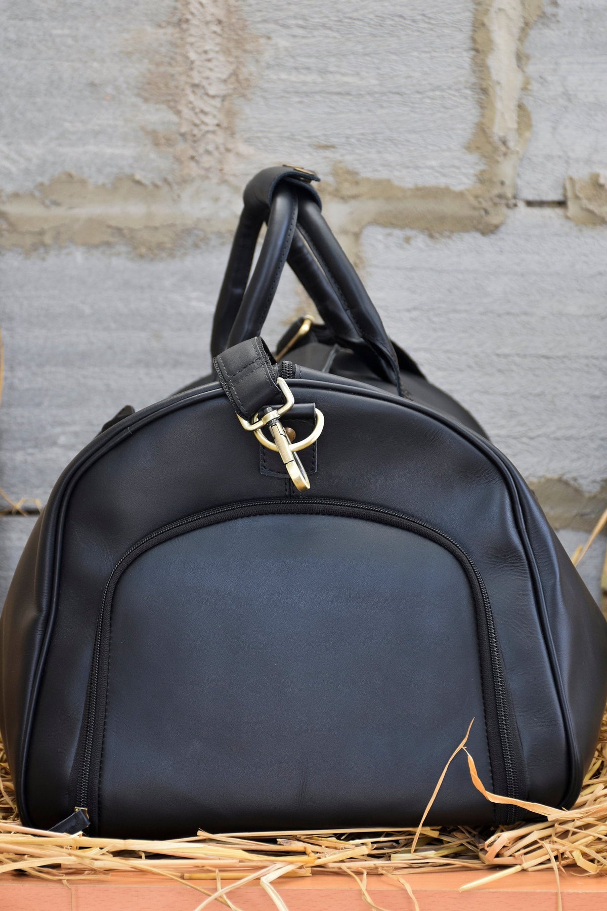 May include: A black duffel bag with a rounded shape, featuring a zippered front pocket and a top handle. The bag has a gold-colored metal clasp and a shoulder strap attachment. The bag is made of a smooth, leather-like material.