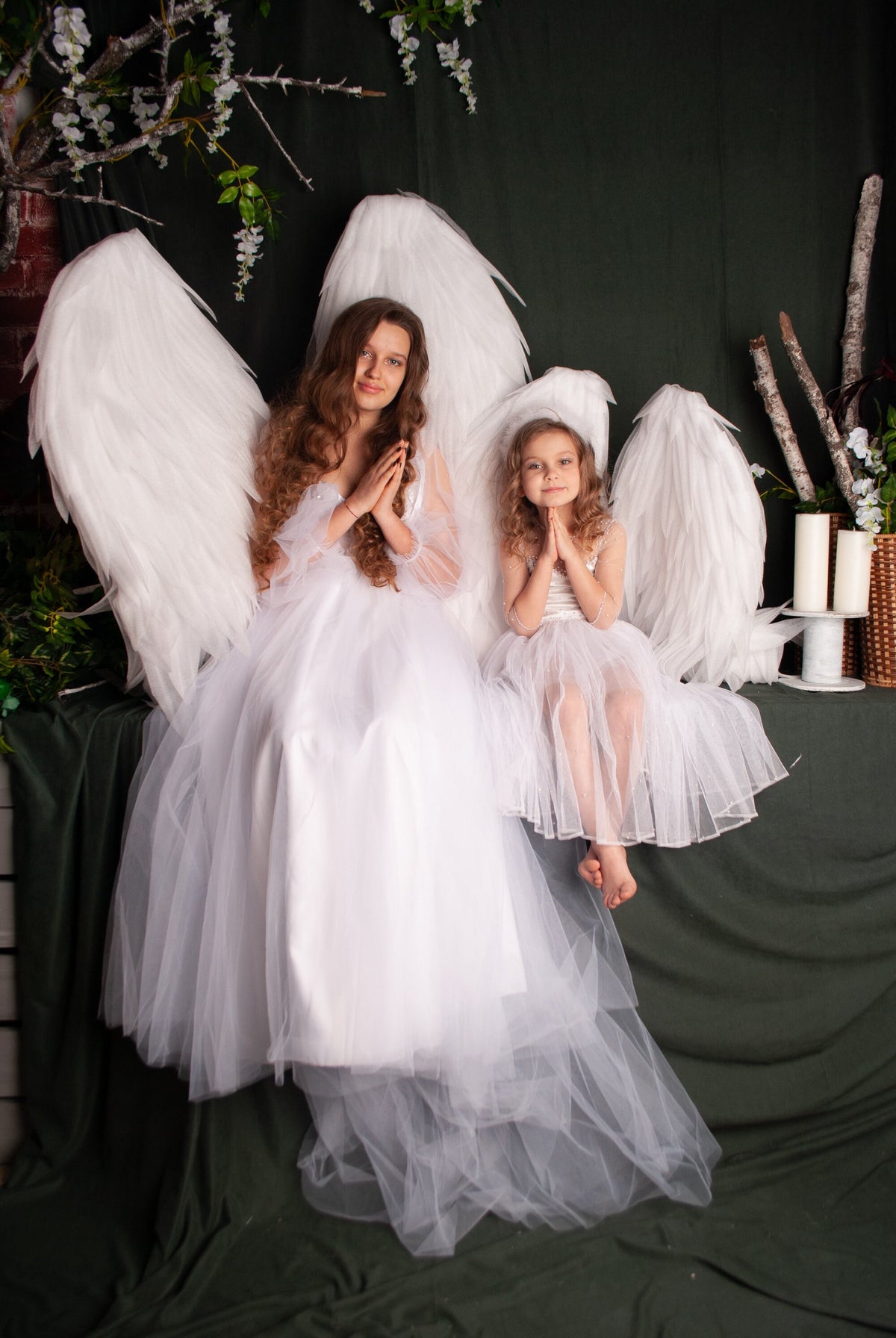 May include: Two girls wearing white dresses and angel wings are posing with their hands clasped in prayer. The girls are sitting on a green surface with white candles and branches in the background.
