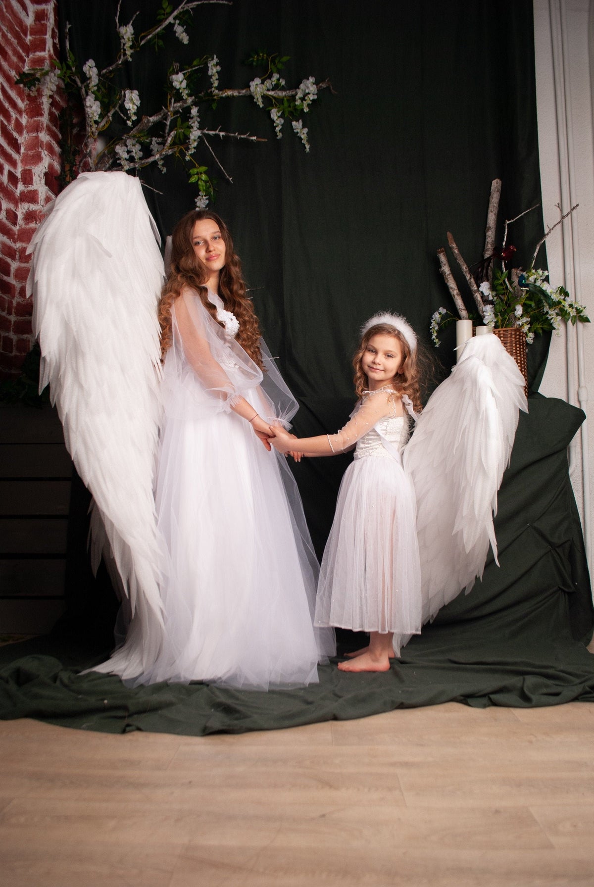 May include: Two girls dressed in white angel costumes with wings, standing on a green surface. The girl in the front is holding the hand of the girl in the back. Both girls are smiling.