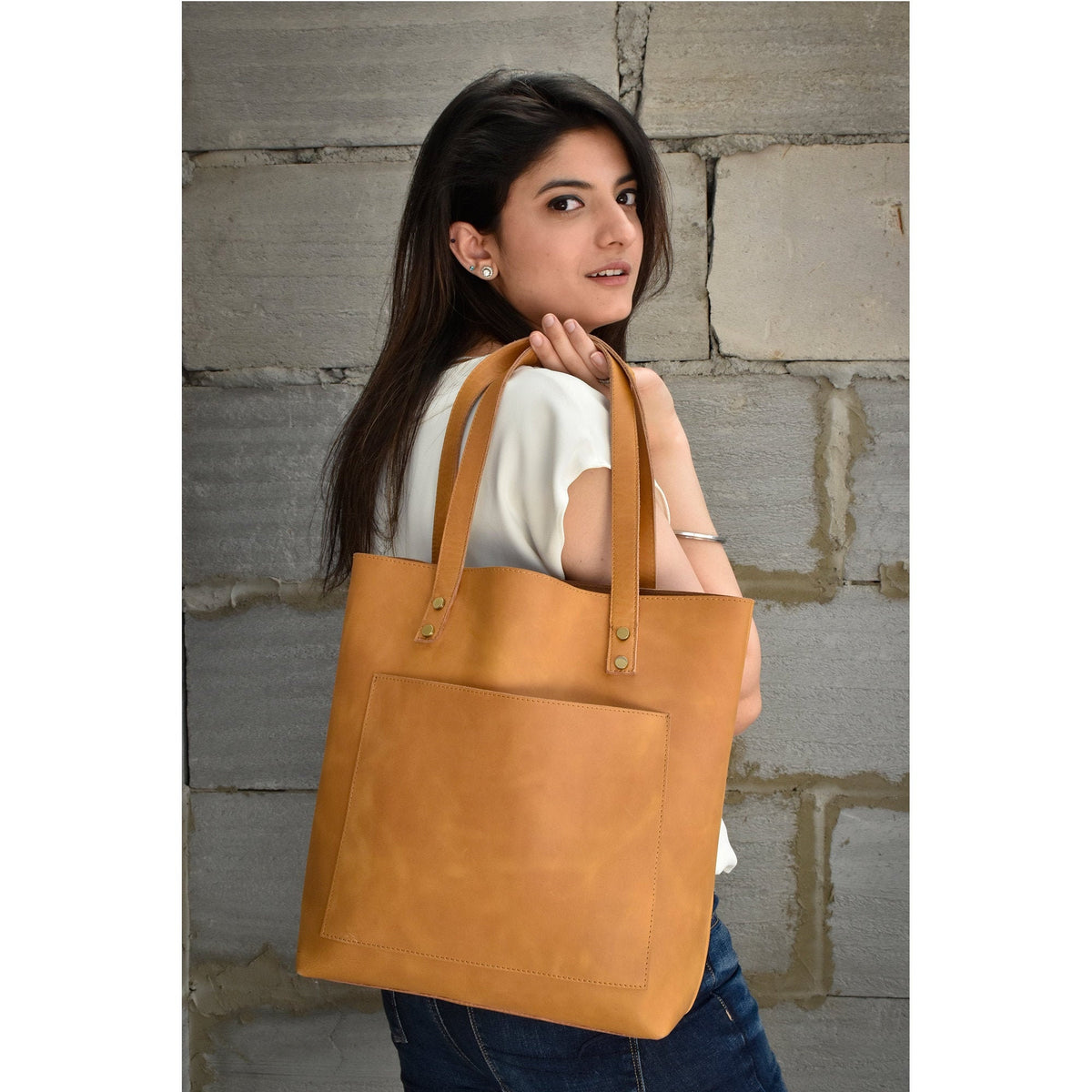 May include: A tan leather tote bag with a front pocket. The bag has brass accents and is worn over the shoulder.