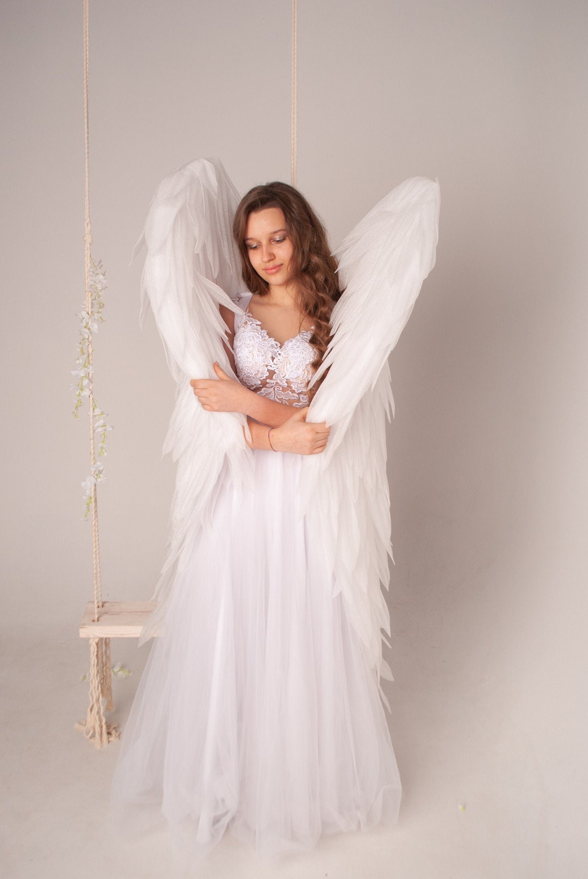 May include: A young woman wearing a white dress and angel wings, standing in front of a white background.
