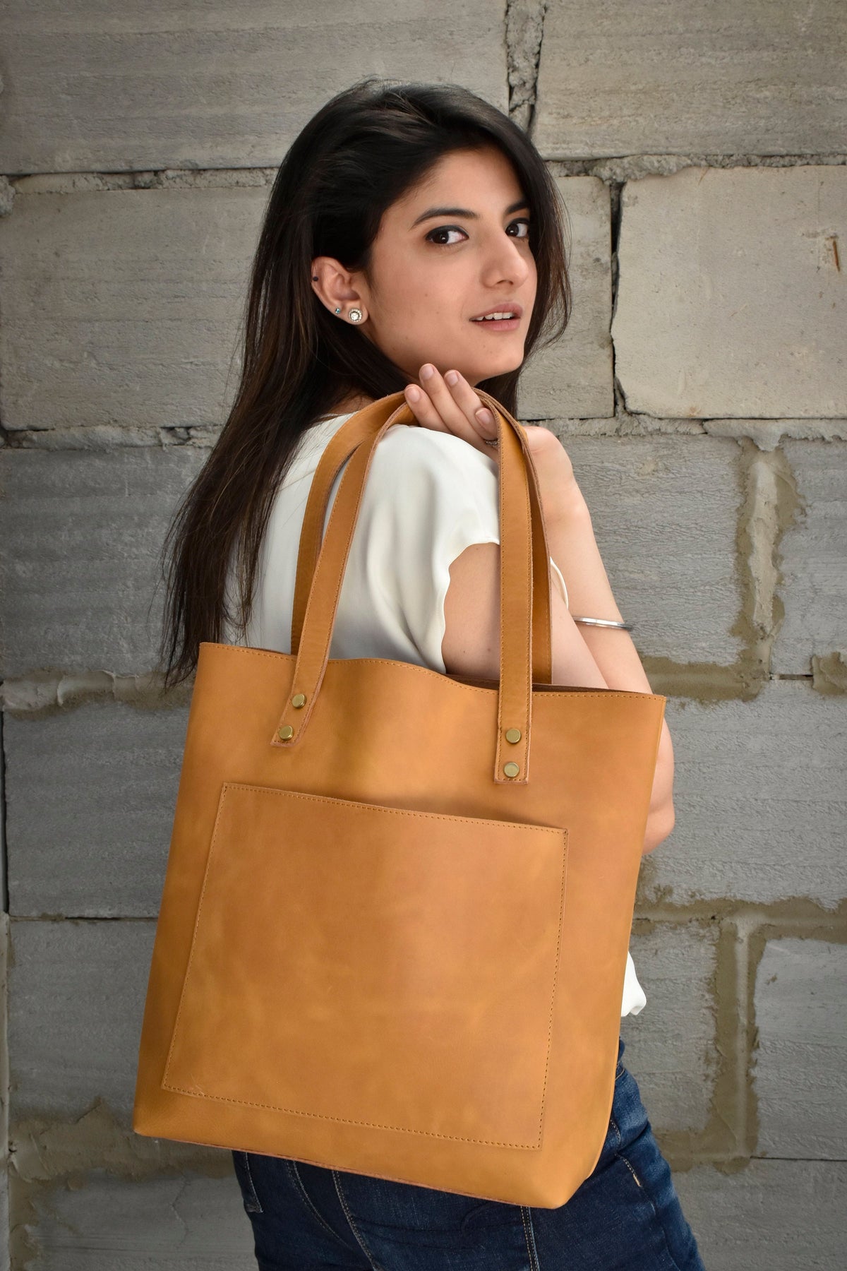 May include: A tan leather tote bag with a front pocket and gold accents. The bag has a wide strap and is being held by a person.