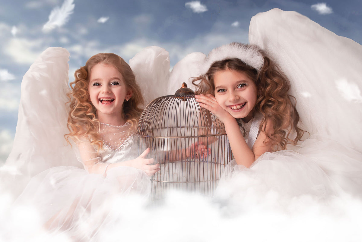May include: Two young girls dressed as angels, smiling and posing with a birdcage. They wear white dresses and feathery wings, set against a cloudy sky background. The image evokes a whimsical, ethereal theme.
