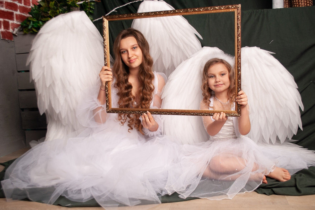 May include: Two girls wearing white angel wings and dresses hold a gold picture frame in front of them. The frame is positioned horizontally and the girls are smiling at the camera.