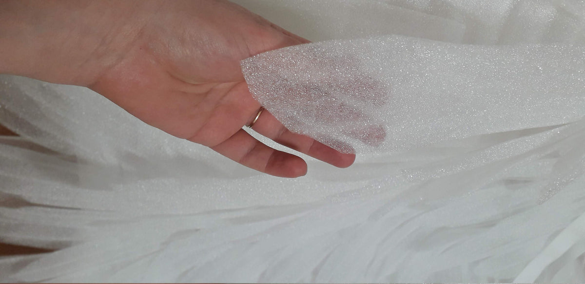 May include: A close-up of white tulle fabric with a glittery, shimmering texture. The fabric is held in a hand, showcasing its delicate and airy quality. The tulle appears to be layered, creating a sense of depth and volume. Ideal for bridal wear or craft projects.