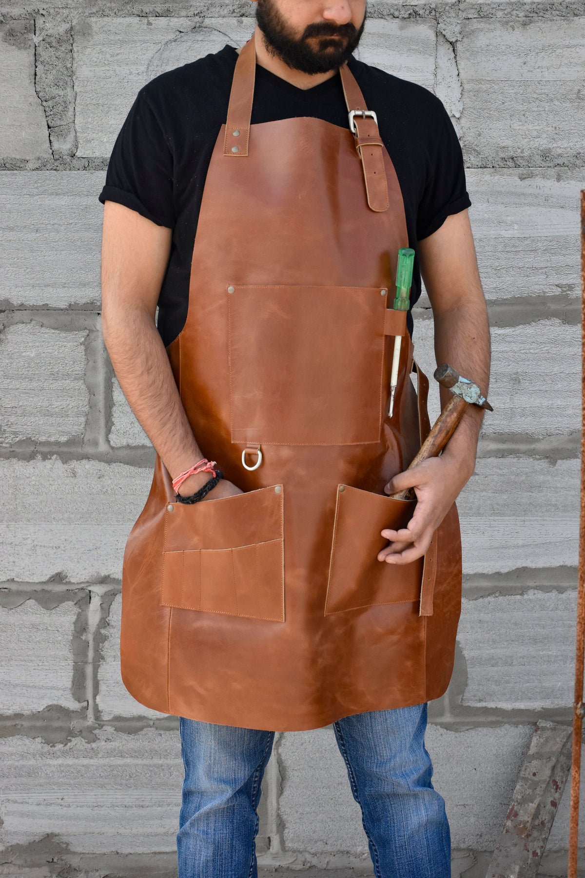 May include: A brown leather apron with multiple pockets and a metal ring. The apron has a strap that goes around the neck and another strap that goes around the waist. The apron is being worn by a person.