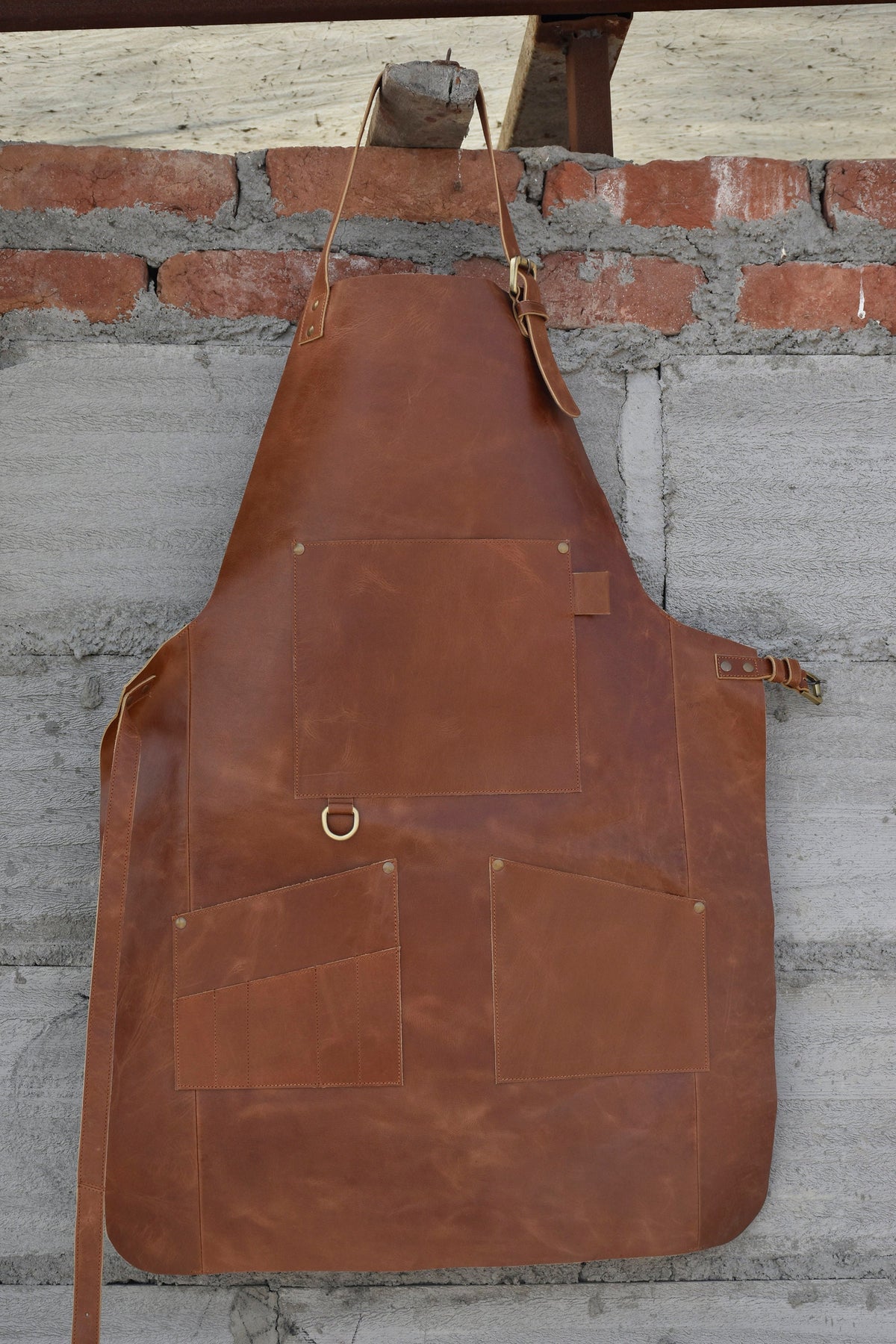 May include: A brown leather apron with a large pocket and two smaller pockets. The apron has a brass buckle and a brass ring.
