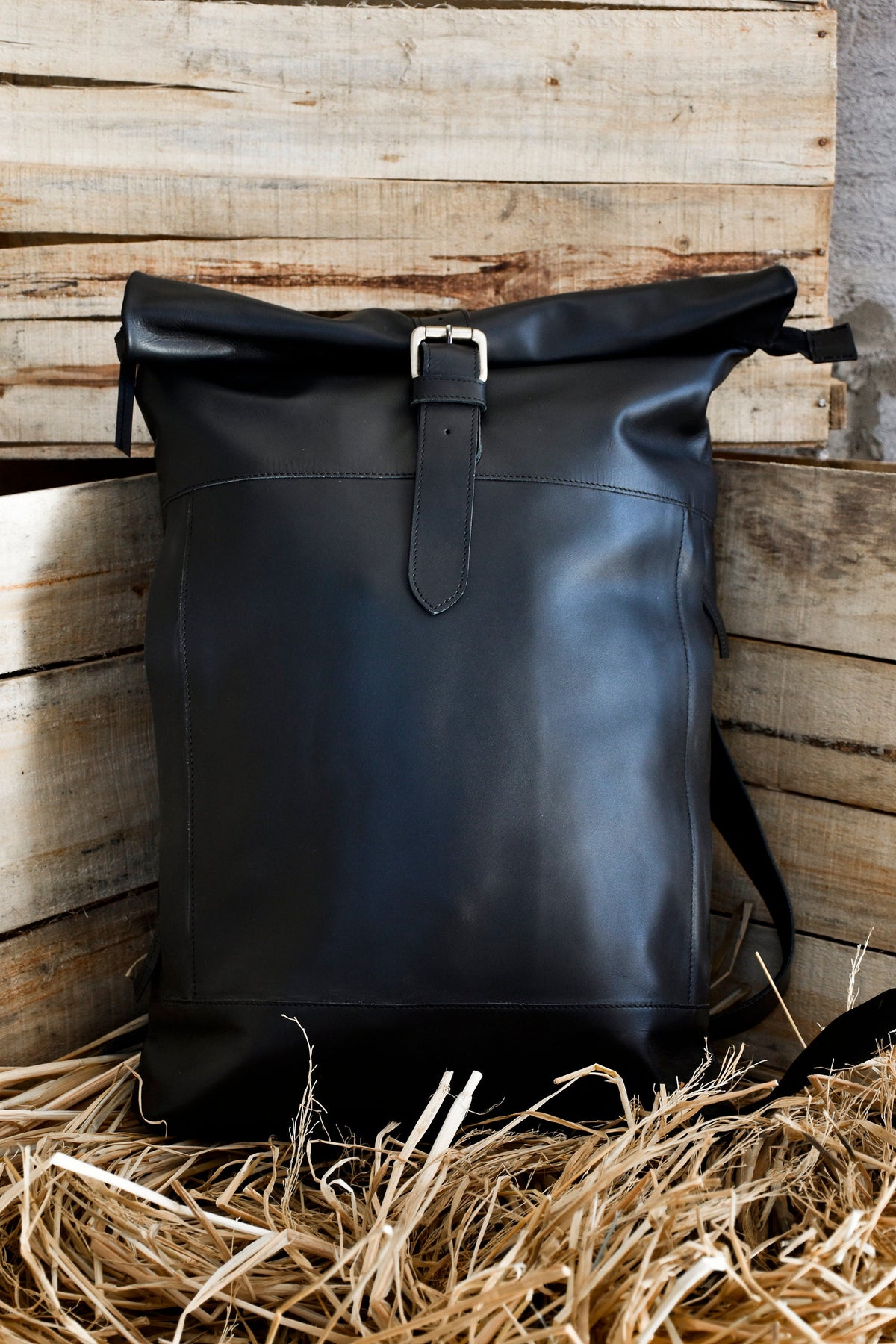May include: A black leather backpack with a roll-top closure and a buckle strap. The backpack is sitting on a bed of straw.