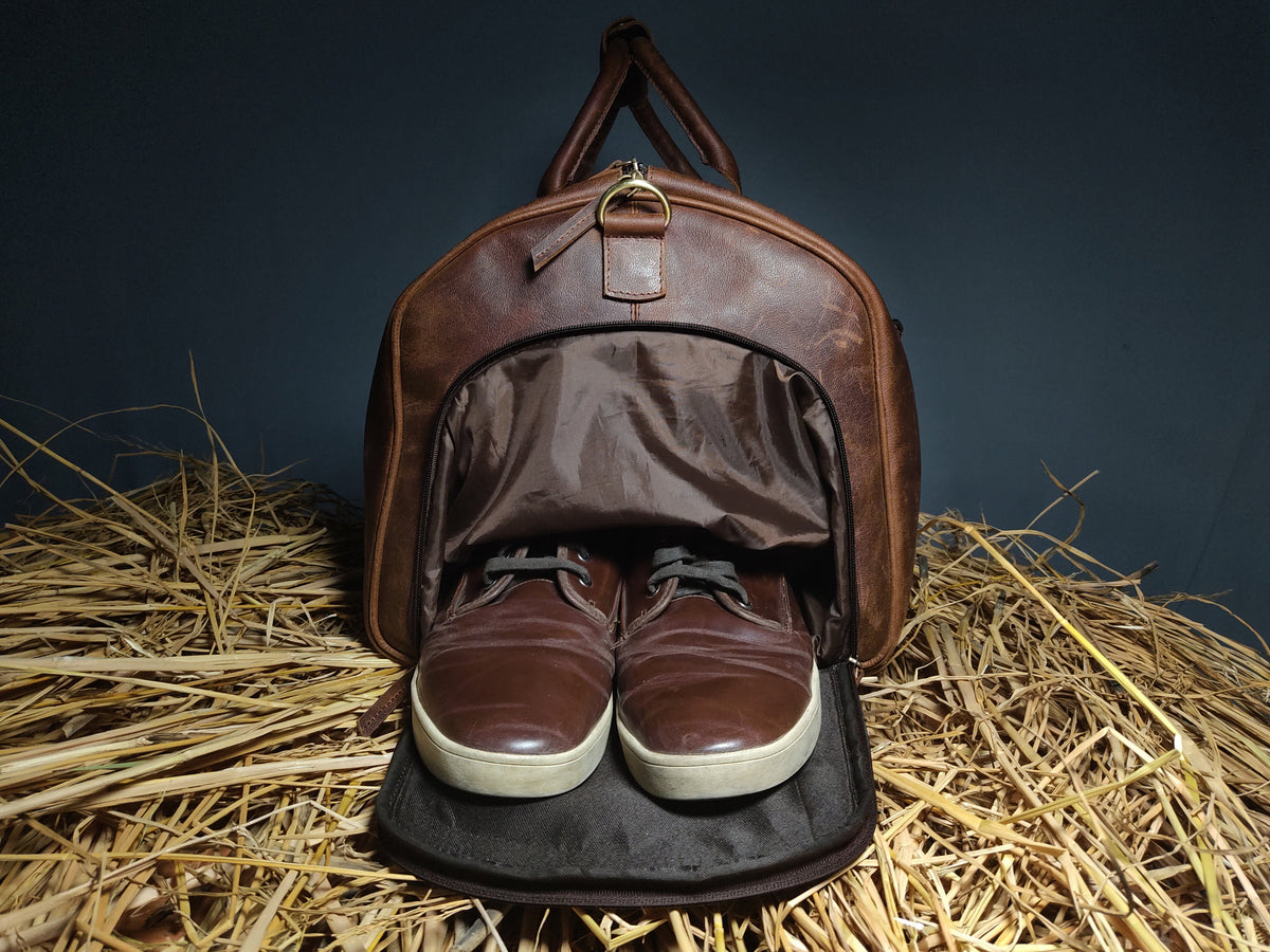 May include: A brown leather duffel bag with a zippered opening, revealing a brown fabric lining. Two brown leather shoes are inside the bag, with the laces tied. The bag is sitting on a bed of straw.