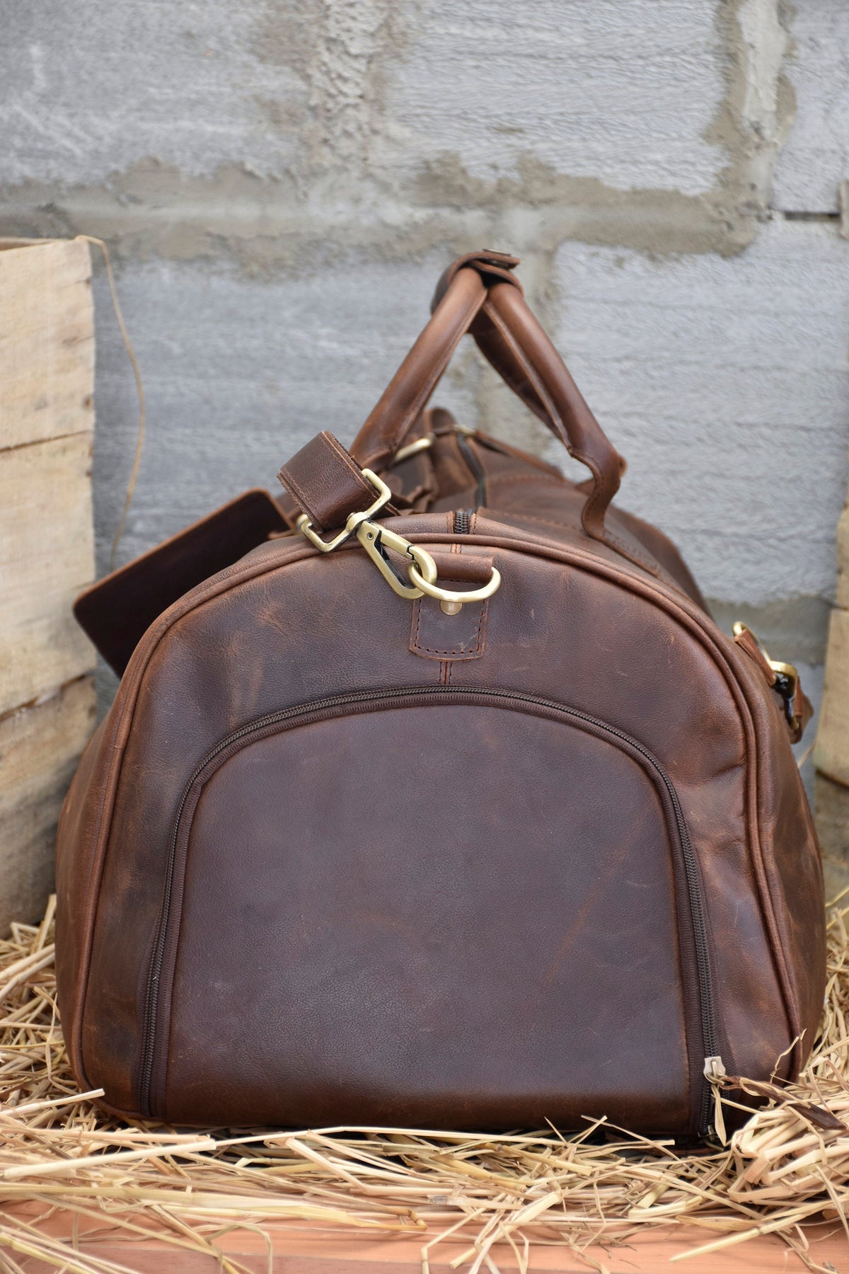 May include: A brown leather duffel bag with a zipper closure and a brass buckle. The bag is sitting on a bed of straw.