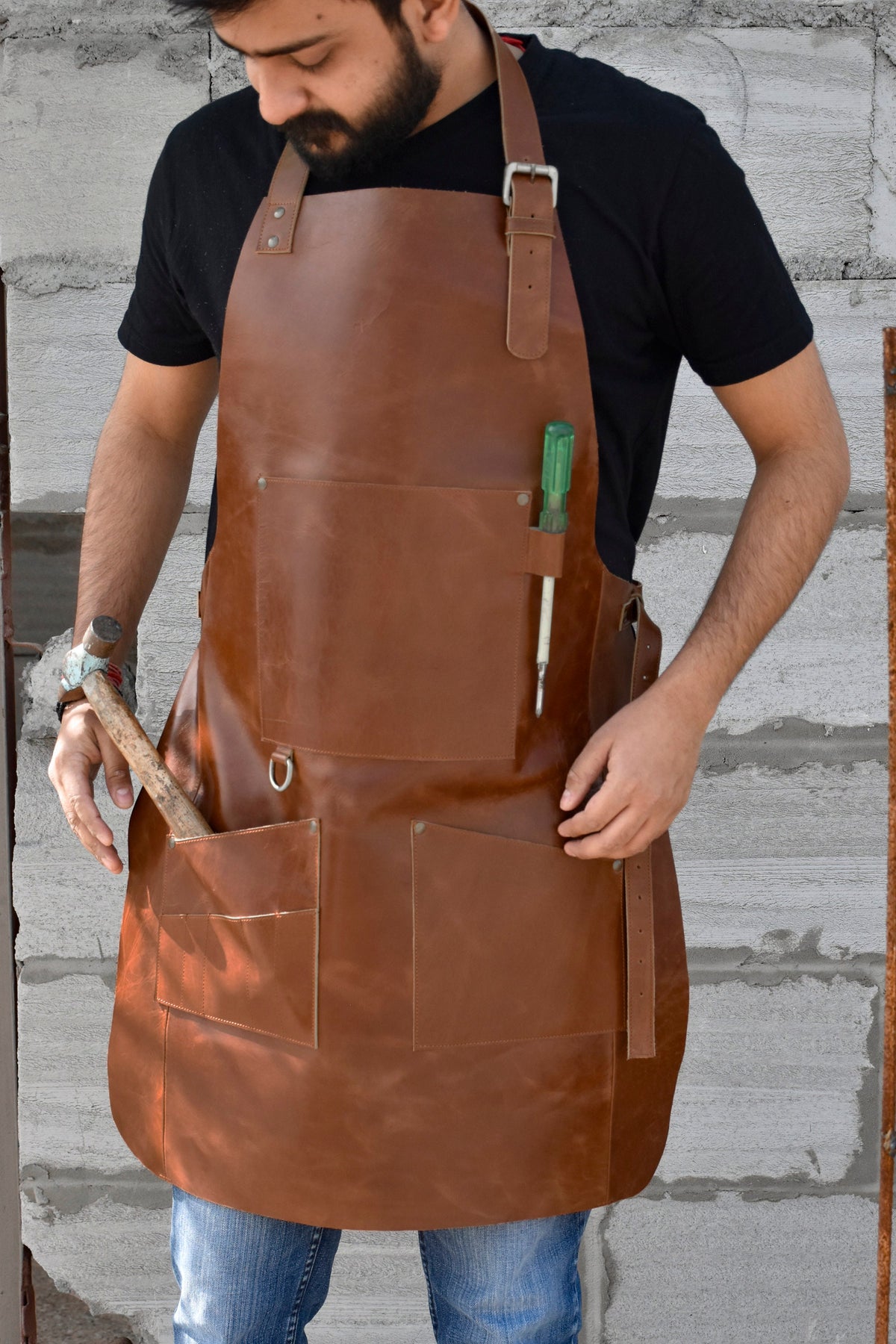 May include: A brown leather apron with multiple pockets and a tool holder. The apron has a buckle strap and is worn by a person.