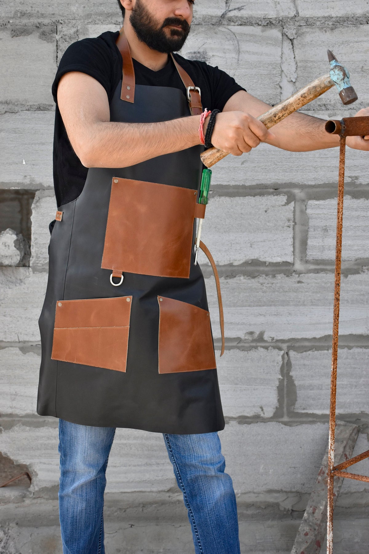 May include: A black leather apron with brown leather pockets and straps. The apron has a tool holder on the side and is being worn by a person holding a hammer.