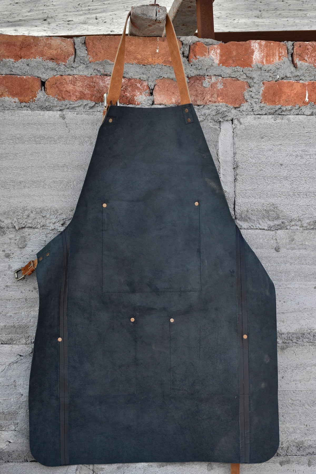 May include: A dark blue leather apron with brown leather straps and a large pocket. The apron has several brass rivets.