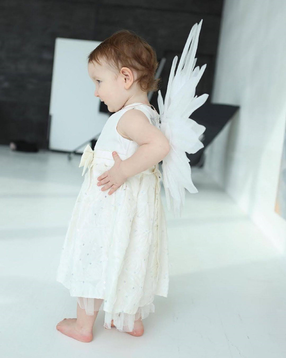 May include: A young child wearing a white dress and angel wings. The child is standing on a white surface and looking to the side.