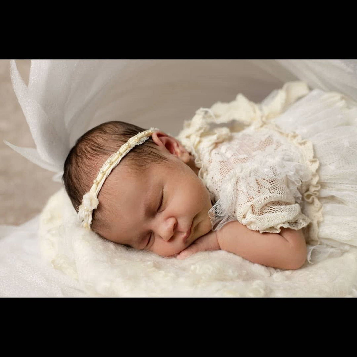 May include: A newborn baby girl sleeping peacefully in a white lace dress with a beaded headband. The baby is lying on a white fluffy surface.
