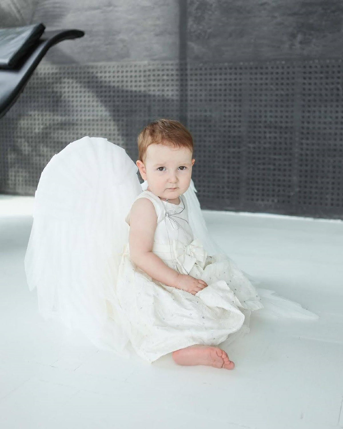 May include: A young child wearing a white dress and angel wings sits on a white surface. The child has red hair and is looking at the camera.