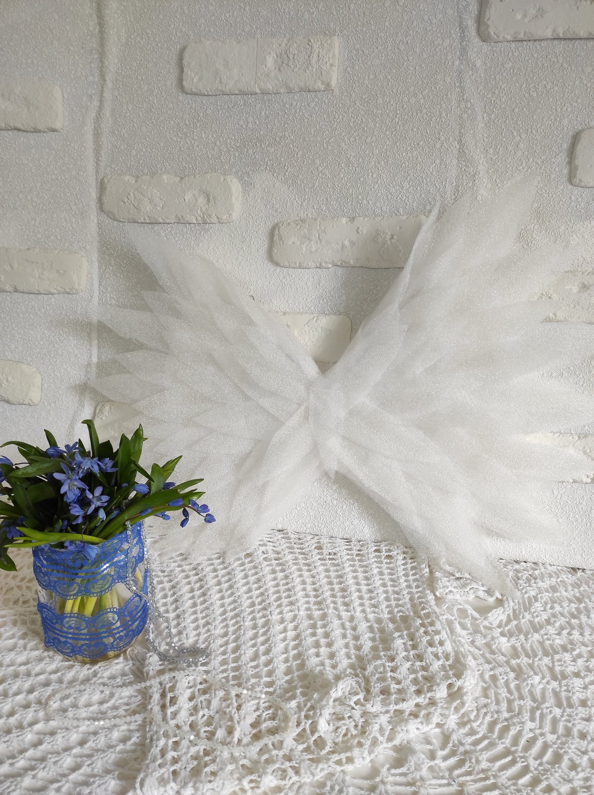 May include: White tulle fabric wings with a delicate, layered design. The wings are displayed against a white brick wall and a white crocheted tablecloth.