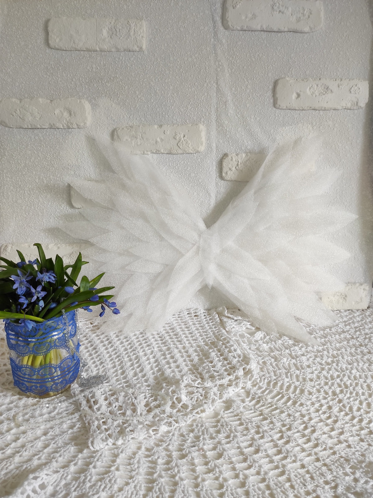 May include: White tulle angel wings against a white brick wall background. A vase with blue flowers sits on a white crocheted tablecloth.