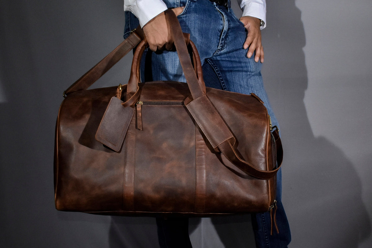 May include: A brown leather duffel bag with a shoulder strap and a luggage tag. The bag has a zipper closure and a handle. The leather has a distressed look.
