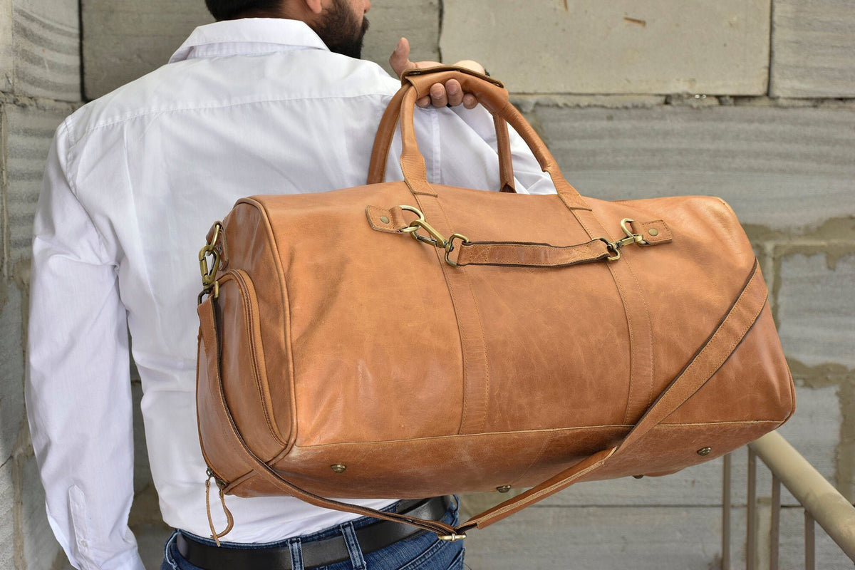 May include: A large, tan leather duffel bag with a shoulder strap and top handles. The bag has brass-colored hardware and a side pocket. The leather appears worn, giving it a vintage look. The bag is designed for travel.