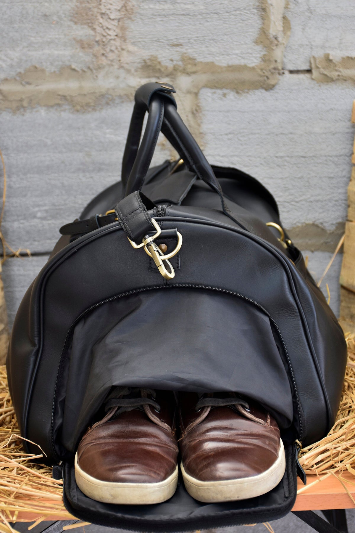 May include: A black leather duffel bag with a top handle and gold-colored hardware. Brown leather shoes with white soles are visible inside the open bag. The bag is set against a textured, light-colored wall.