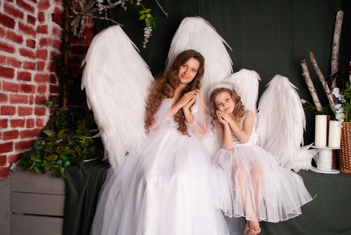 May include: Two girls wearing white angel wings and dresses, with long hair, are posing with their hands clasped together. They are sitting in front of a brick wall and green foliage.