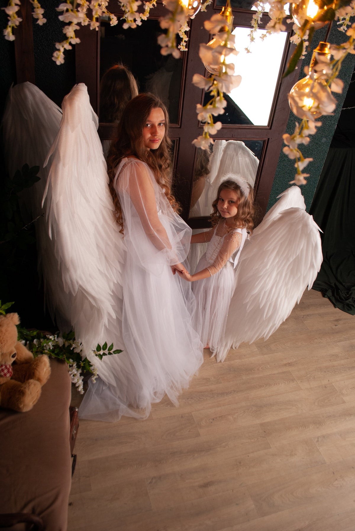 May include: Two girls wearing white dresses and angel wings stand in front of a mirror. The girl in the front is holding the hand of the girl behind her. The girls are smiling and looking at the camera.