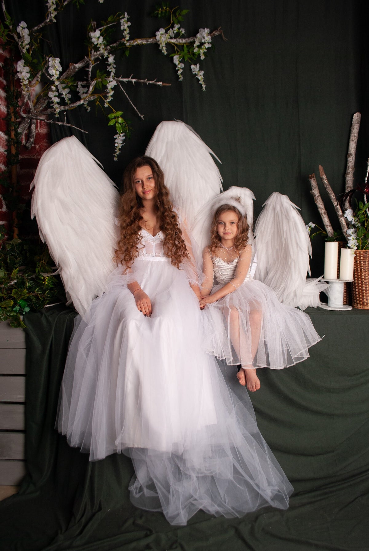 May include: Two girls wearing white dresses and angel wings pose for a photo. The girl in the front is holding a white feather. The girls are sitting on a green surface with a wooden crate and white candles in the background.