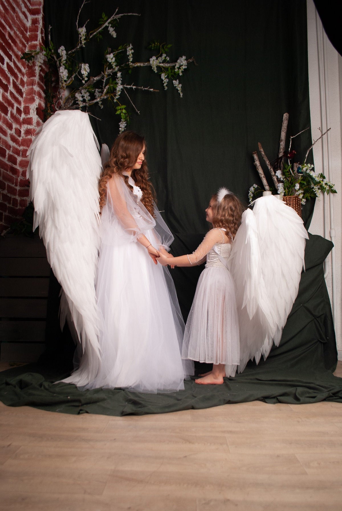 May include: Two people dressed as angels, wearing white dresses and large white wings, stand facing each other and holding hands. The background is a dark green fabric with white flowers.