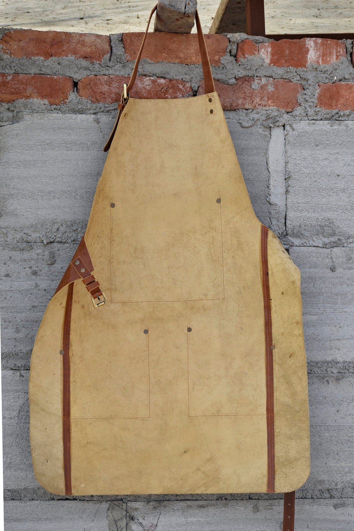 May include: A tan leather apron with brown leather straps and a buckle. The apron has multiple pockets and is hanging on a brick wall.