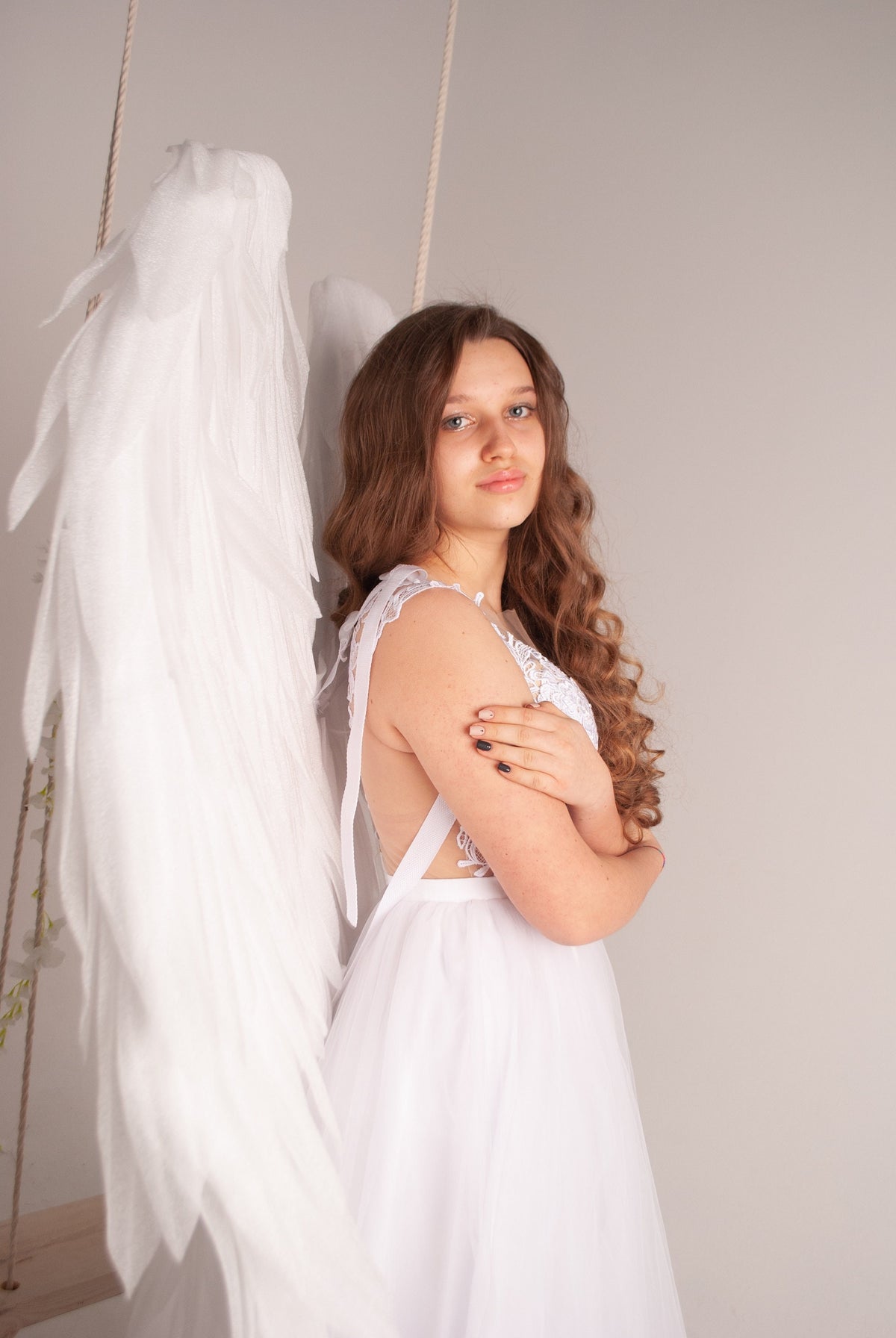 May include: A young woman with long brown hair wearing a white dress and white angel wings. She is standing in front of a white background.