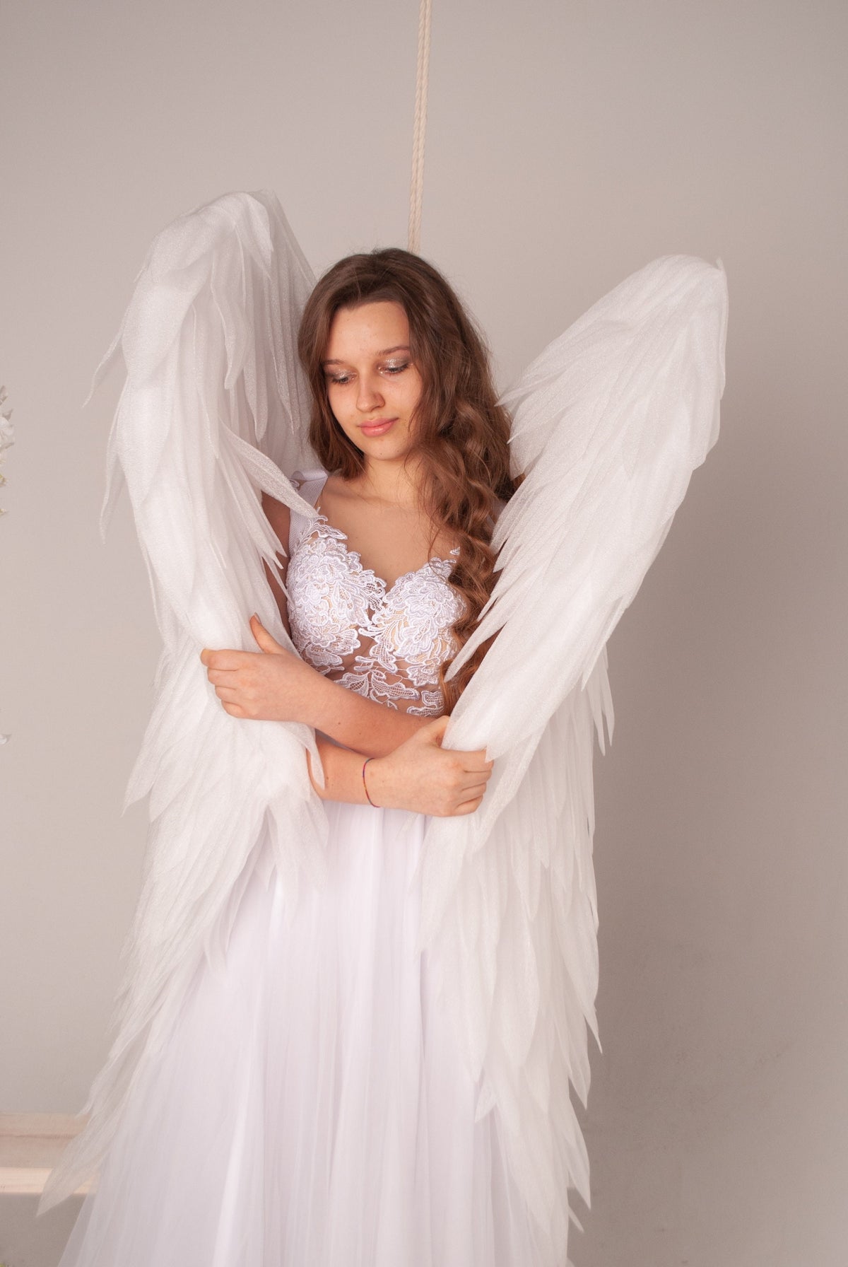 May include: A young woman wearing a white lace dress and large white angel wings. She is looking down and holding the wings close to her body.