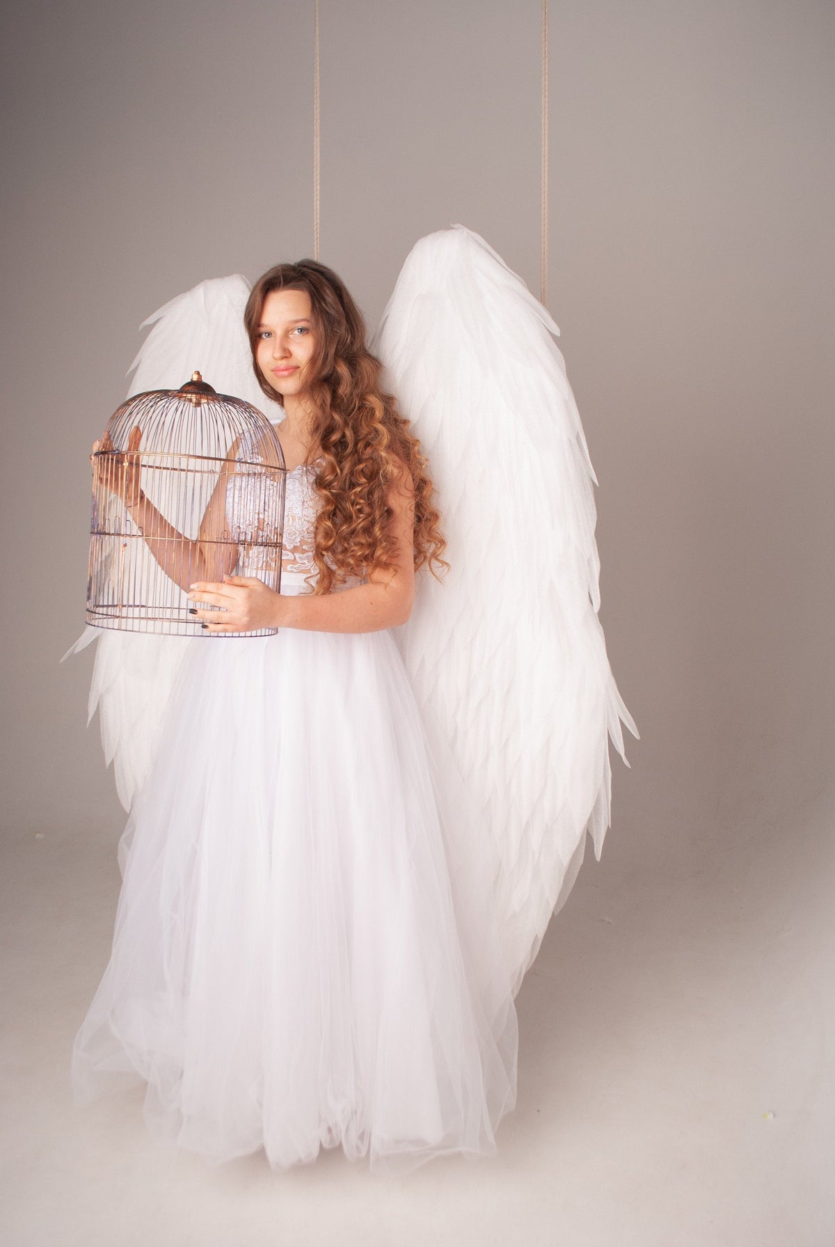 May include: A young woman wearing a white dress and large white angel wings holds a birdcage in front of her. The cage is made of metal and has a rounded top.