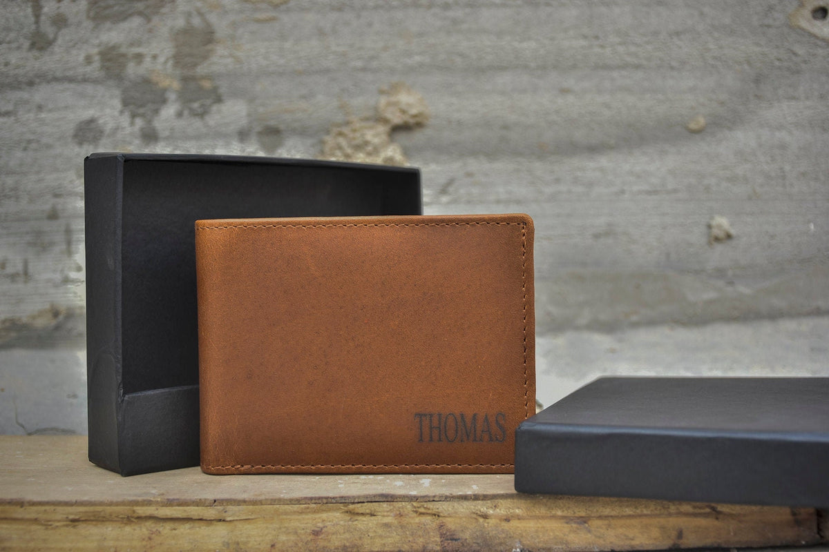 May include: A brown leather wallet with the name "Thomas" embossed on the front. The wallet is sitting on a wooden surface next to two black boxes.