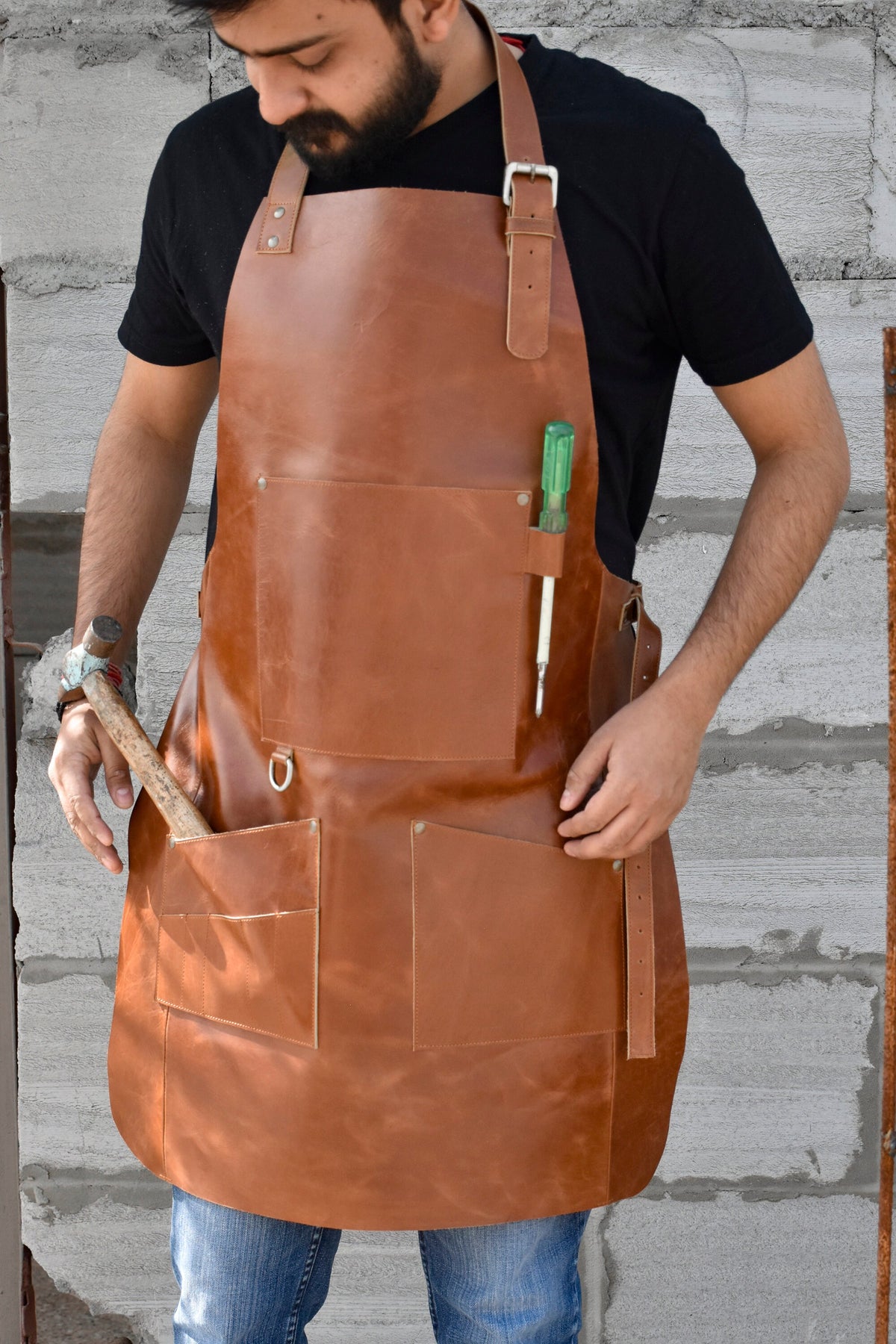 May include: A brown leather apron with multiple pockets and a tool holder. The apron has a buckle strap and is worn by a person.