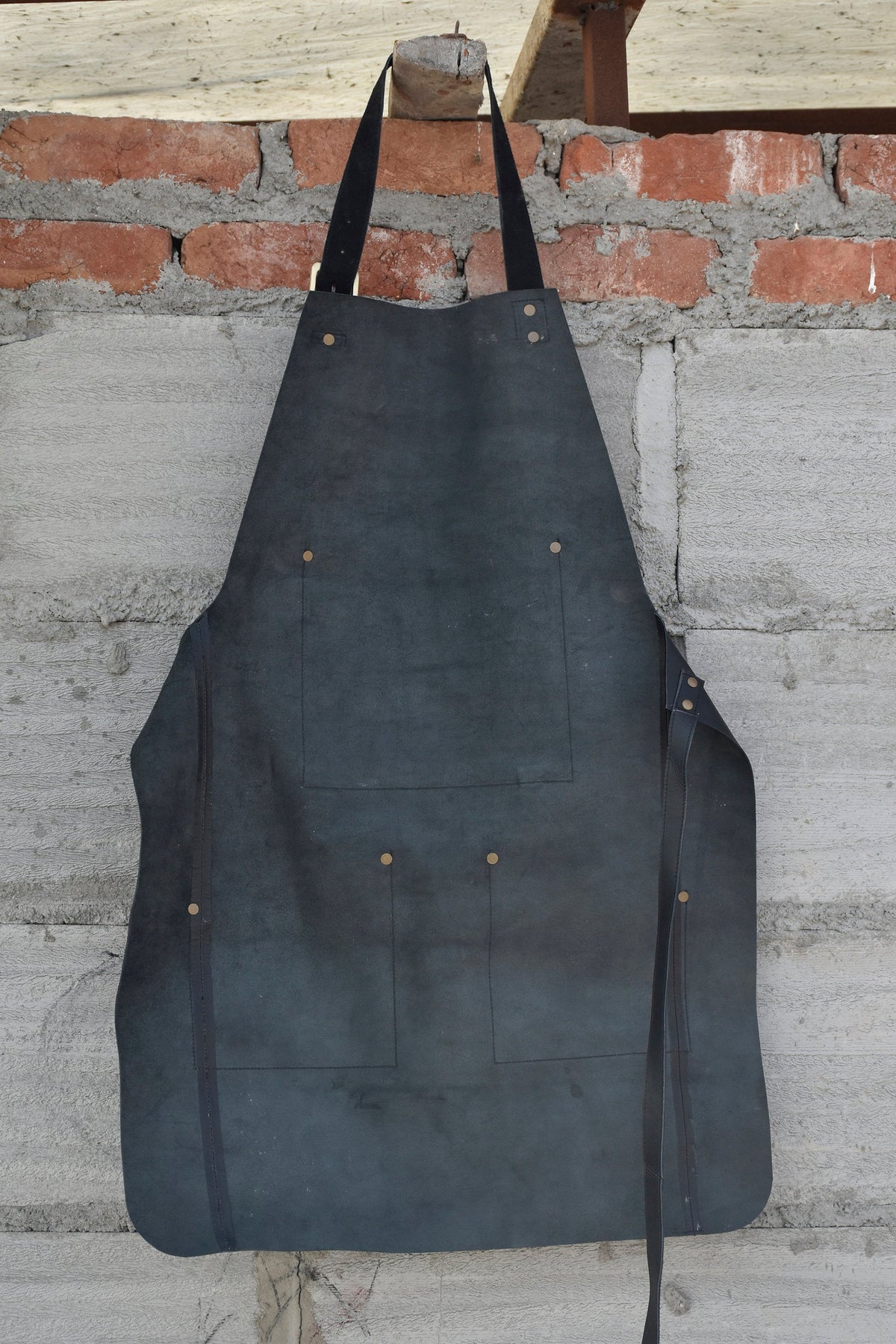 May include: A black leather apron with multiple pockets and straps. The apron is hanging on a brick wall.