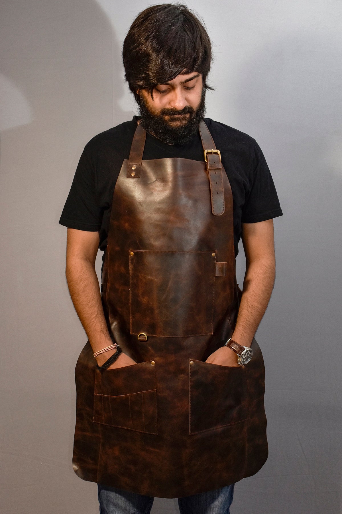 May include: A brown leather apron with multiple pockets and a brass buckle. The apron is worn by a person with a beard and a black t-shirt.