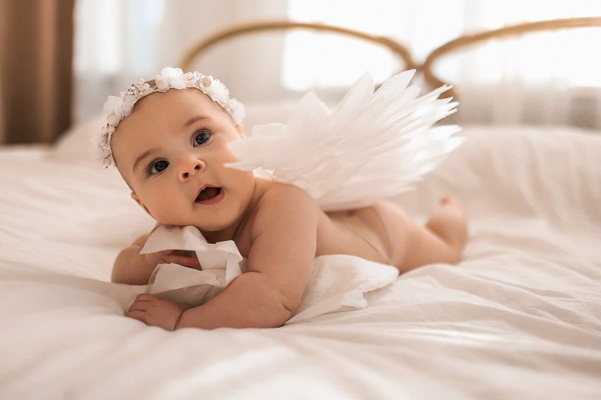May include: A baby wearing a white feathered angel costume and a white flower crown lies on a white bed, looking at the camera with a surprised expression.