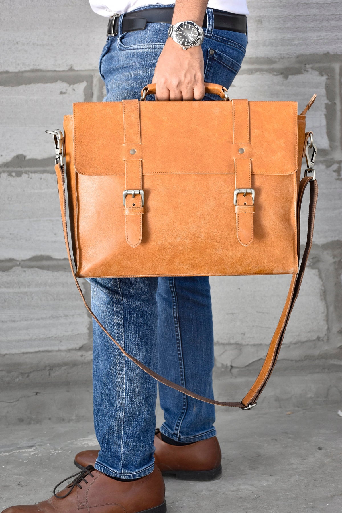 May include: A brown leather briefcase with a long strap and two buckles. The briefcase has a flap with a buckle closure.